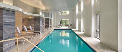 An indoor swimming pool with clear blue water, surrounded by lounge chairs and a sunbed area.