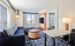 A hotel room with a sofa, ottoman, dining table, chair, TV, lamp, and large windows with curtains. - Embassy Suites by Hilton Washington D.C. – Convention Center (Washington)