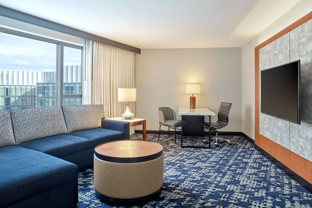 A hotel room with a large window, a sofa, a round coffee table, a desk with chairs, and a flat-screen TV mounted on the wall.