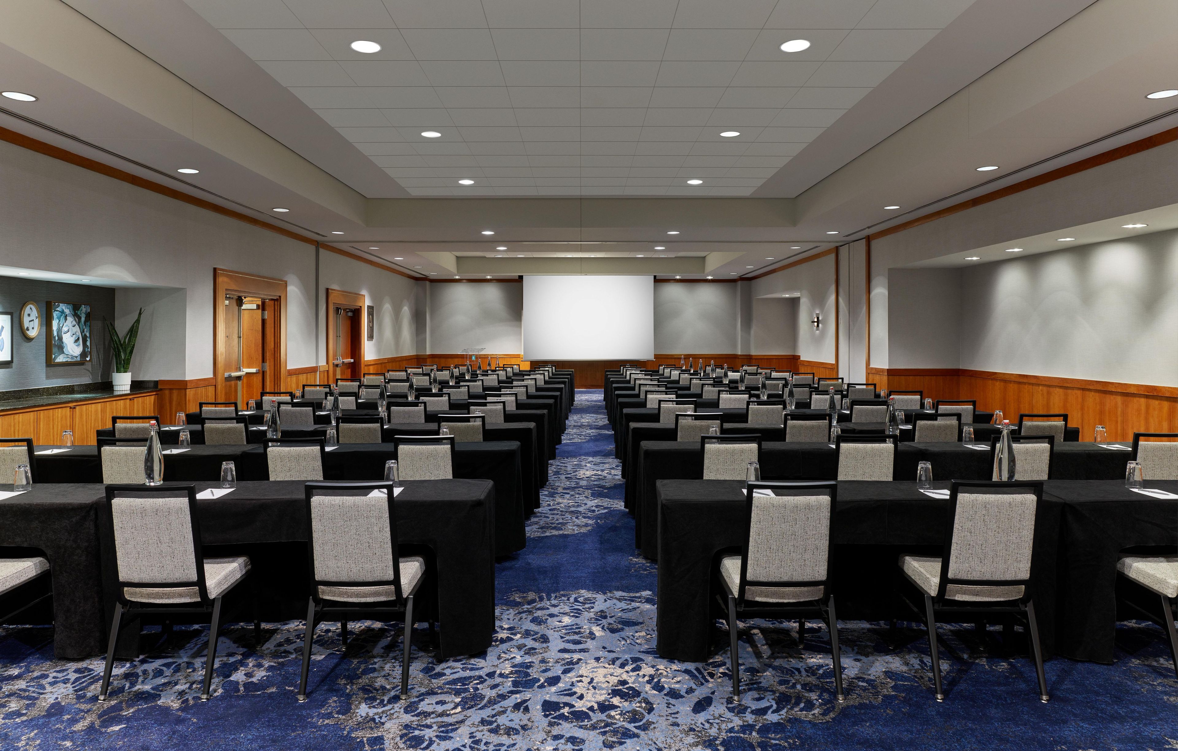 A conference room with rows of tables and chairs, a large screen, and a carpeted floor.