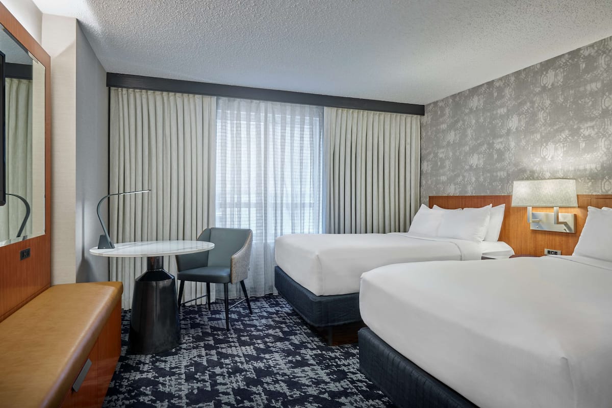 A hotel room with two beds, a small table with a chair, a bench, a TV, and a large window with curtains.