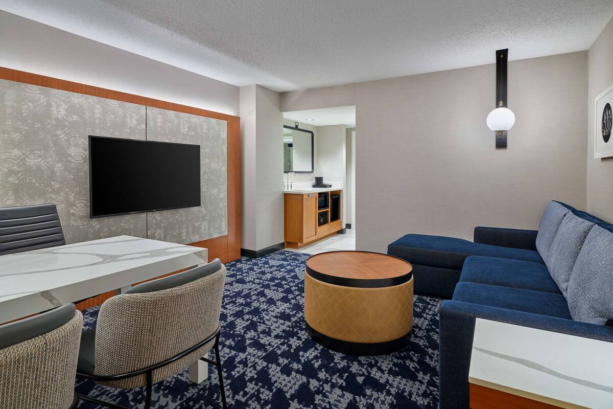A modern hotel room with a flat-screen TV, a dining table with chairs, a sofa, a round ottoman, and a bathroom visible through an open door.