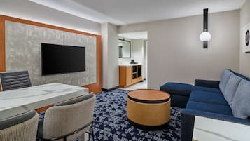 A modern hotel room with a flat-screen TV, a dining table with chairs, a sofa, a round ottoman, and a bathroom visible through an open door.