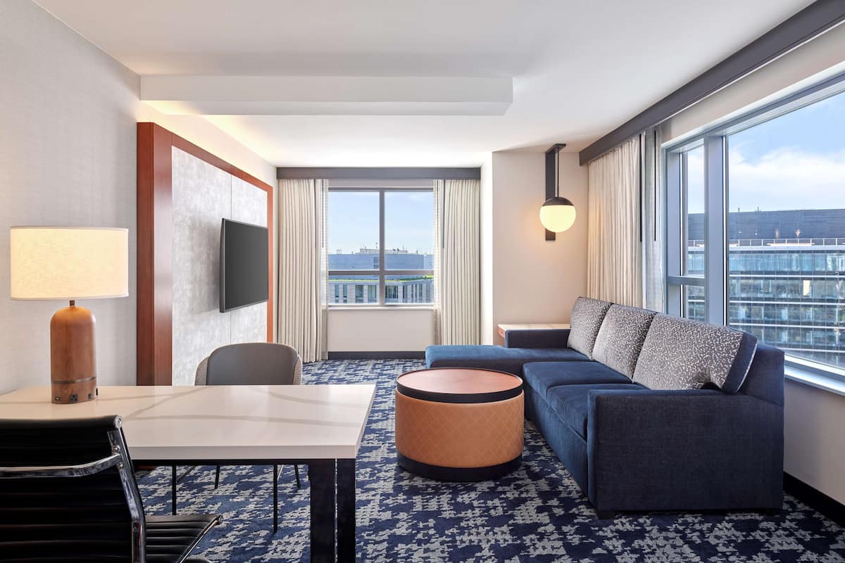 A hotel room with a large window, a sofa, a round ottoman, a table, and a TV on the wall.