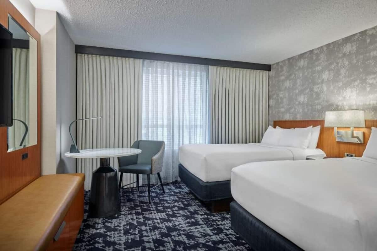 A hotel room with two beds, a small table with a chair, a bench, a TV, and a large window with curtains.