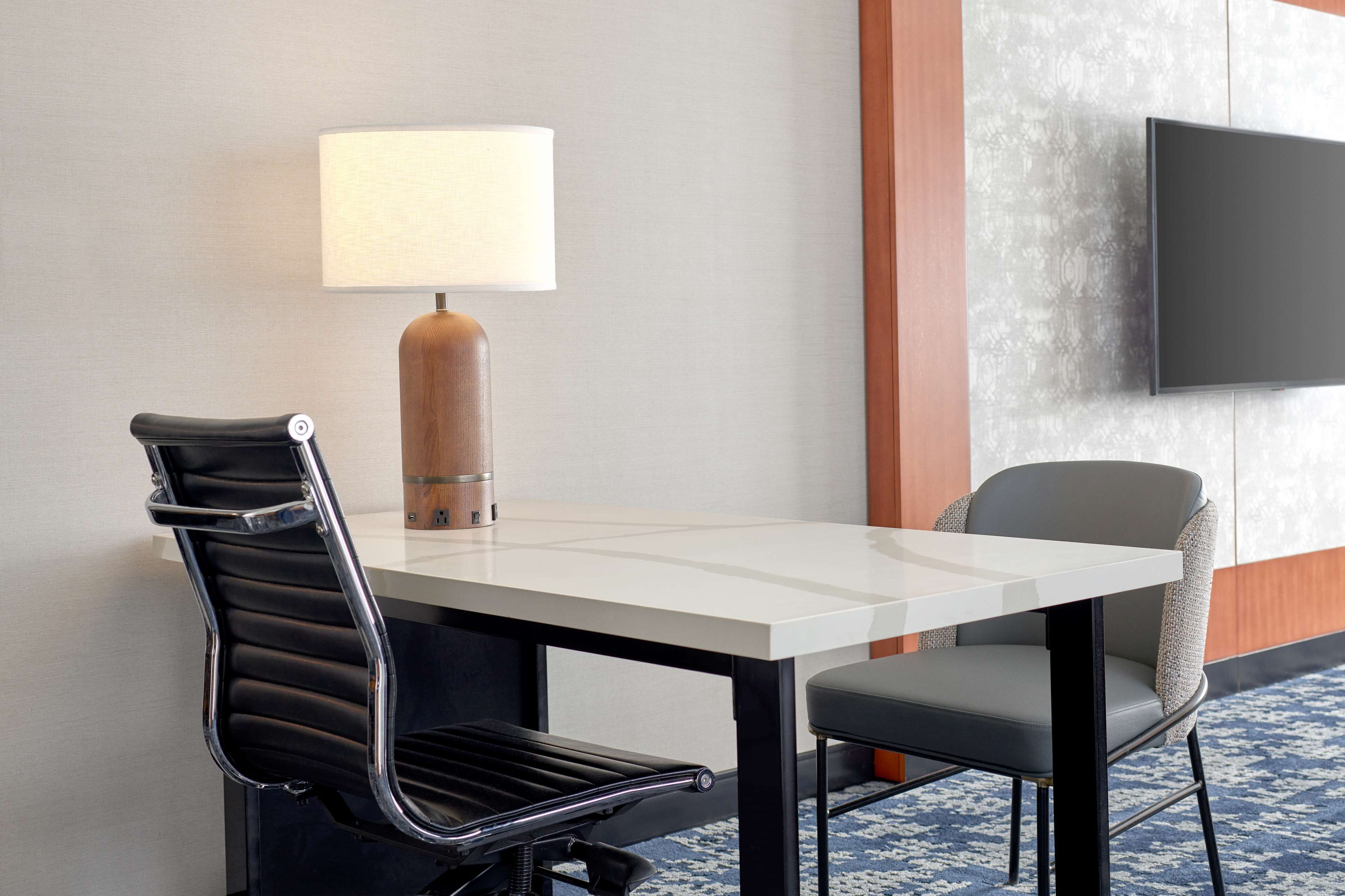 A hotel room with a modern desk, two chairs, a lamp, and a flat-screen TV mounted on the wall.
