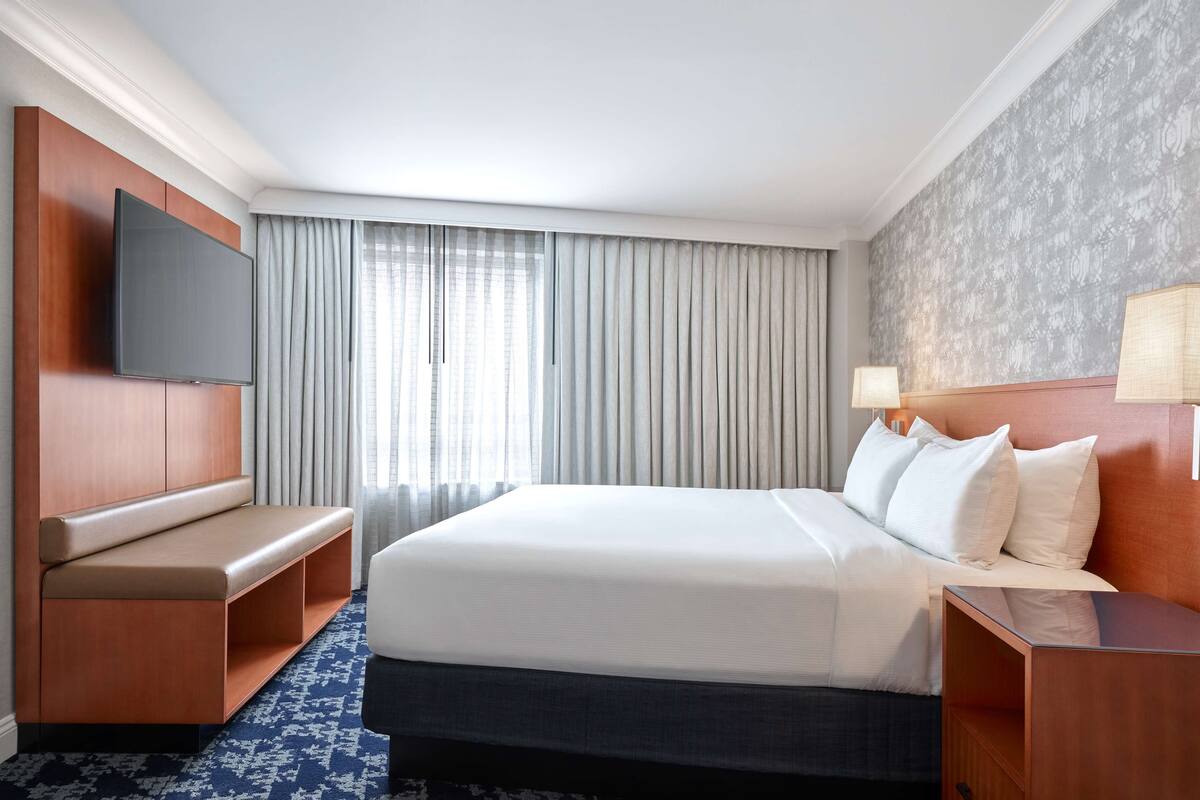 A hotel room with a large bed, a flat-screen TV mounted on the wall, a bench with storage, a bedside table with a lamp, and a patterned carpet.