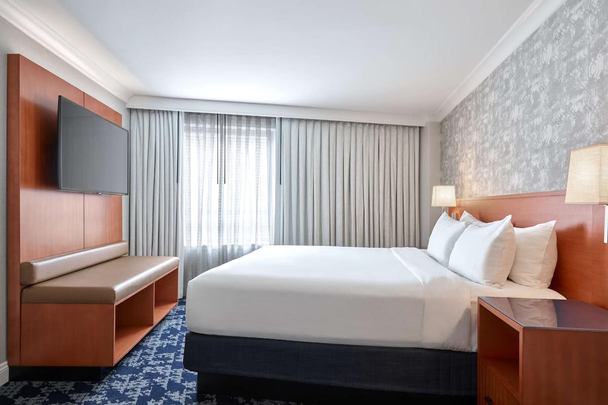 Una habitación de hotel con una cama grande, un televisor de pantalla plana montado en la pared, un banco con espacio de almacenamiento, una mesita de noche con lámpara y una alfombra estampada.