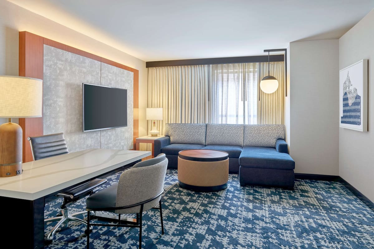 A hotel room with a large flat-screen TV, a sofa, a round ottoman, a desk with chairs, and a patterned carpet.