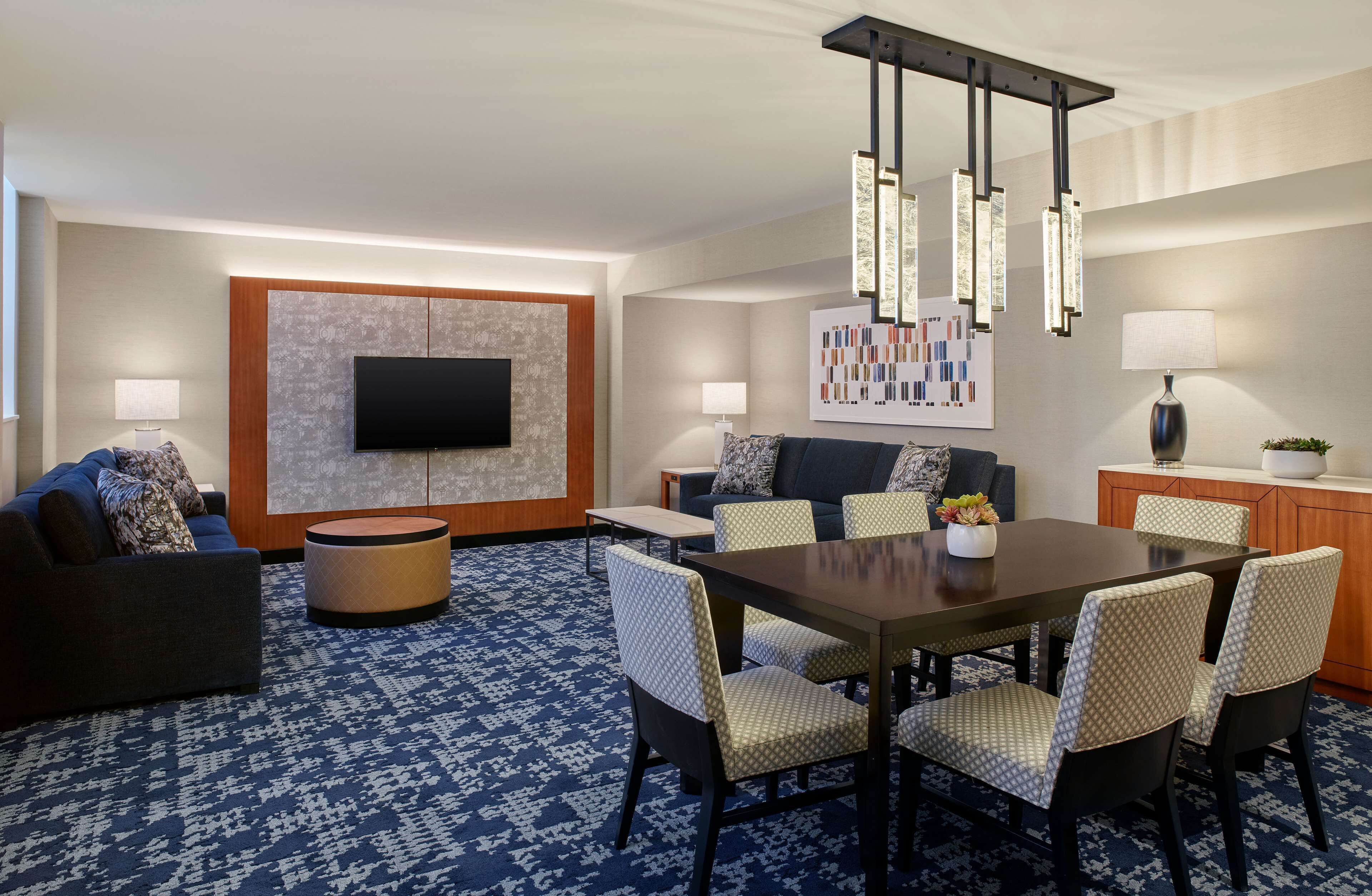 A modern hotel room with a flat-screen TV, a dining area with a table and chairs, a sofa, and a patterned carpet.