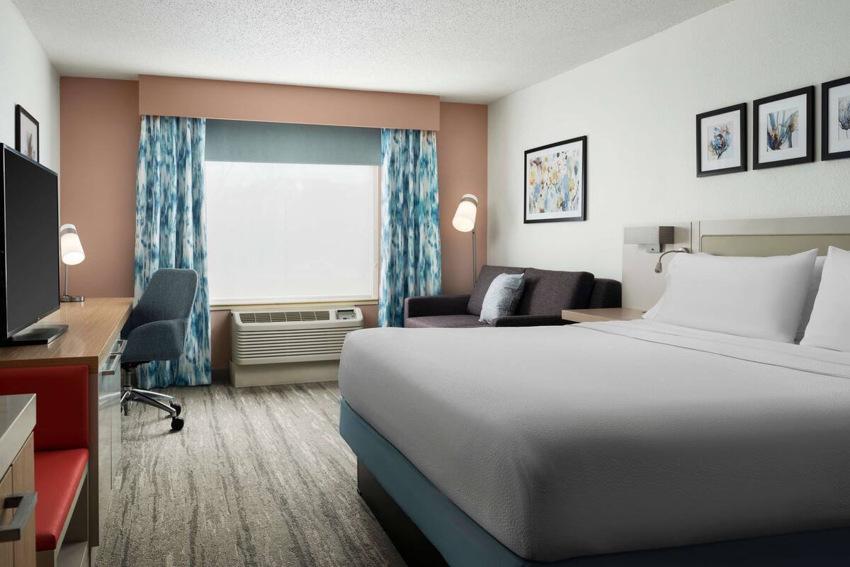 A hotel room with a large bed, a desk with a computer, a chair, a TV, and a window with curtains.