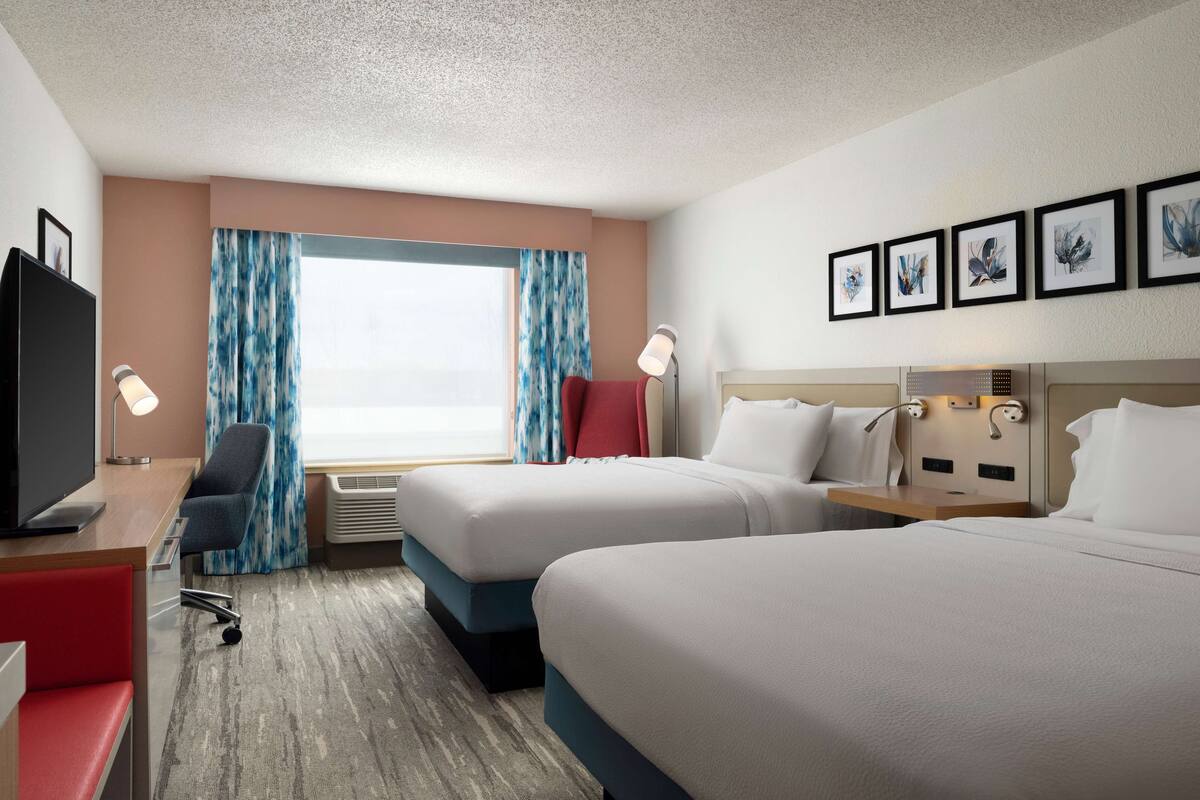 A hotel room with two beds, a desk with a TV, a chair, and a window with curtains.