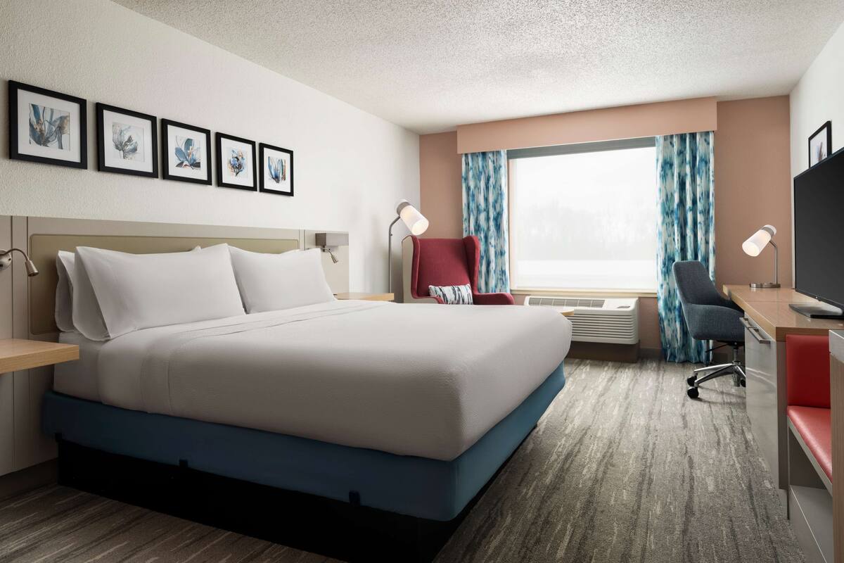 A hotel room with a large bed, a desk, a chair, a TV, and a window with curtains.