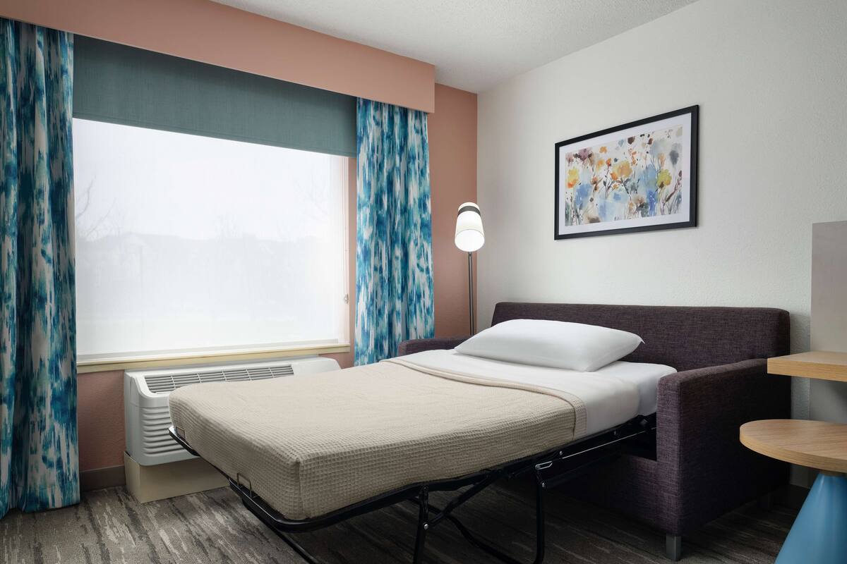 A hotel room with a bed, a bedside lamp, a window with curtains, a radiator, and a painting on the wall.