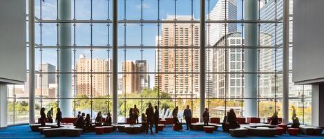 A spacious indoor area with a high ceiling and large windows, offering views of tall buildings and a clear sky.