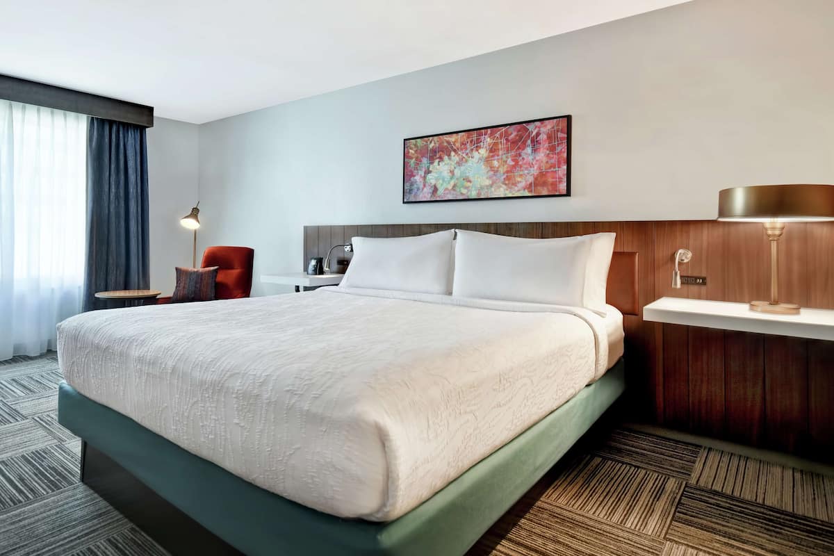 A hotel room with a large bed, a wooden headboard, a desk with a chair, a lamp, and a painting on the wall.