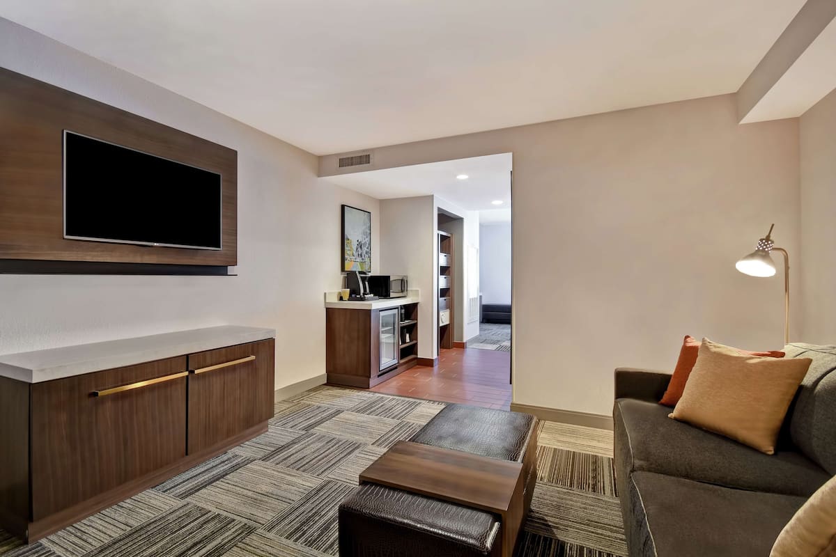 A modern living room with a flat-screen TV, a sofa, a coffee table, and a kitchenette in the background.
