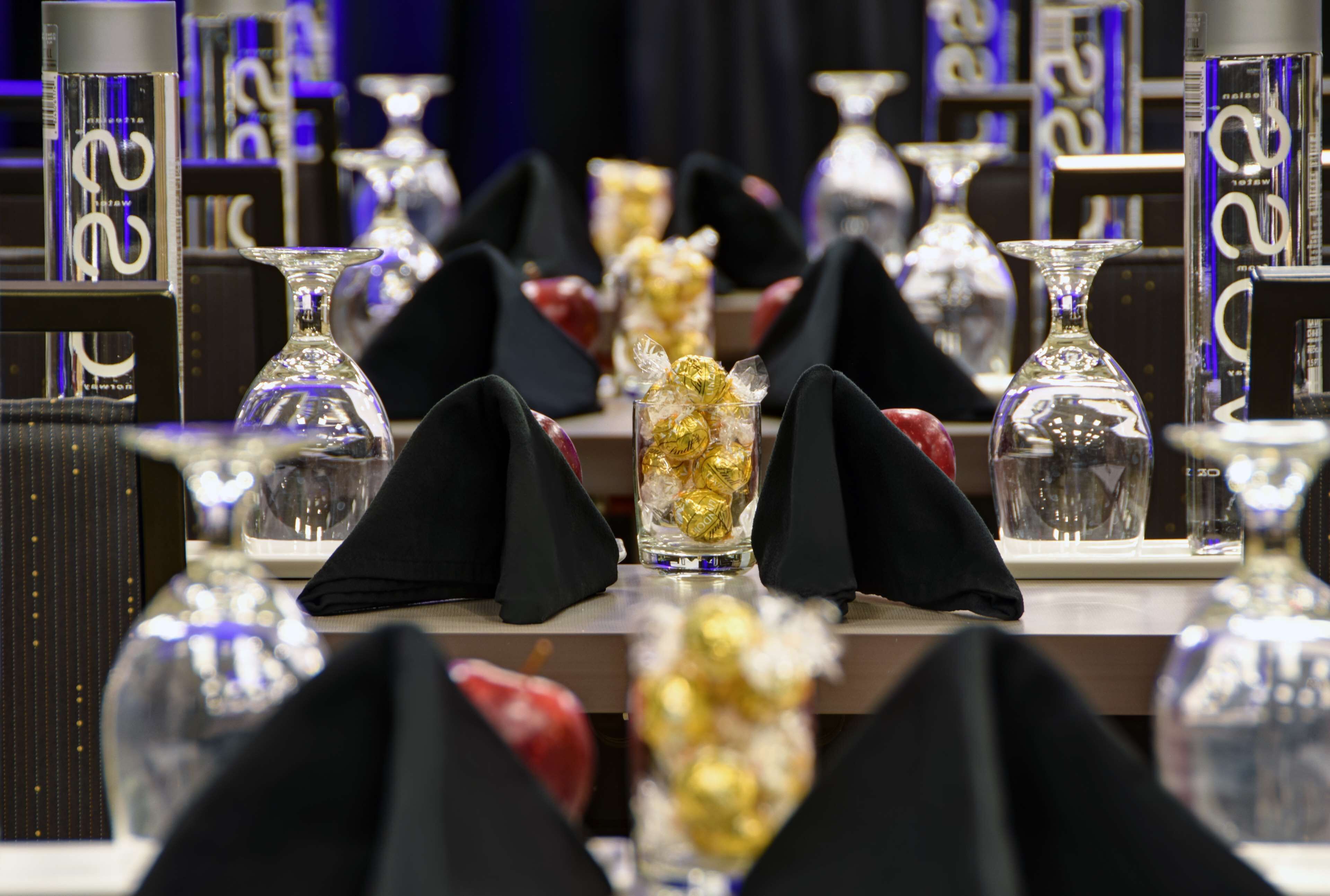 Une table dressée avec de beaux verres transparents et des chocolats emballés dans du papier doré, entourée de serviettes noires et de boîtes à l’effigie de la marque.