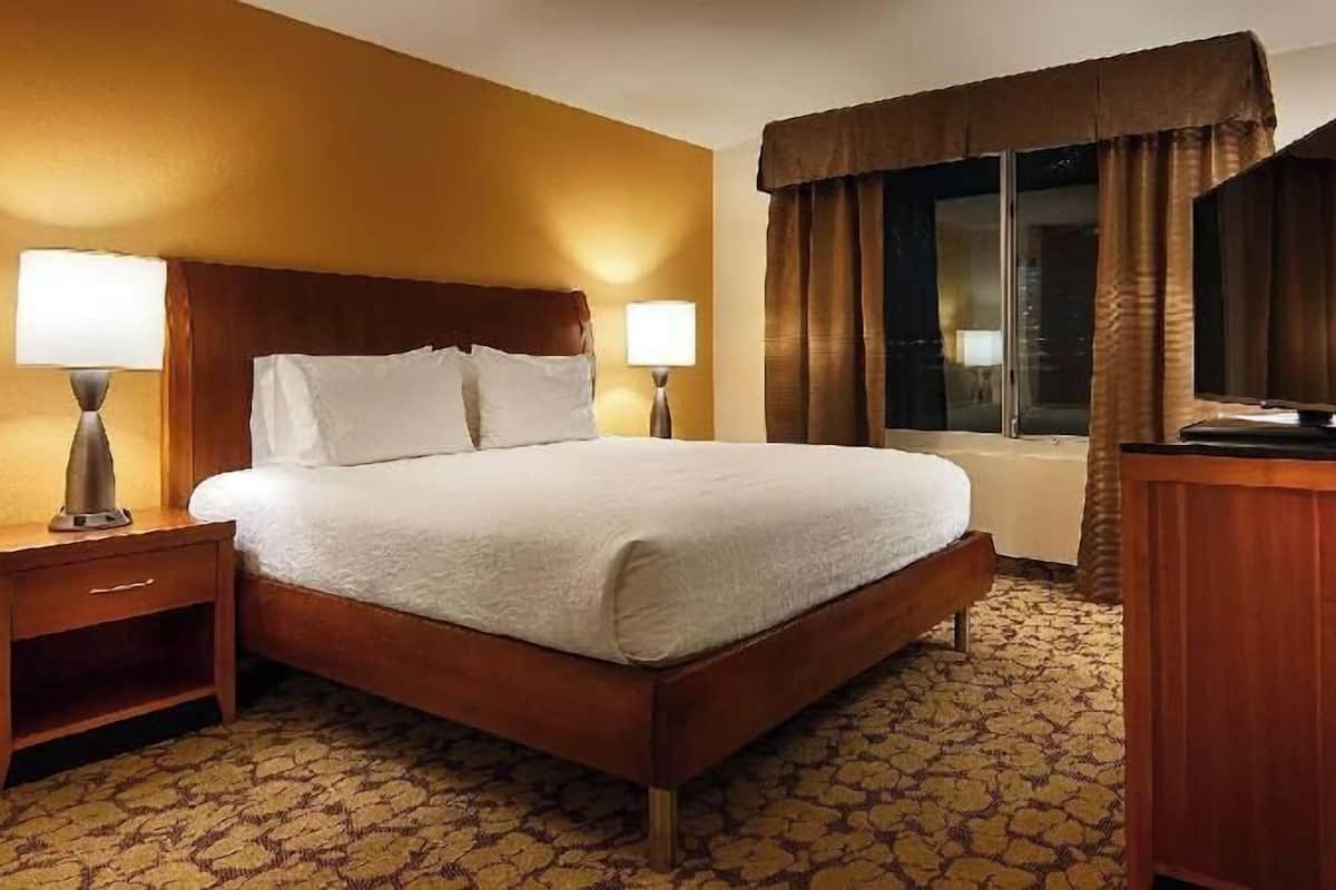 A hotel room with a wooden bed, two bedside lamps, a wooden desk, a television, and a window with curtains.