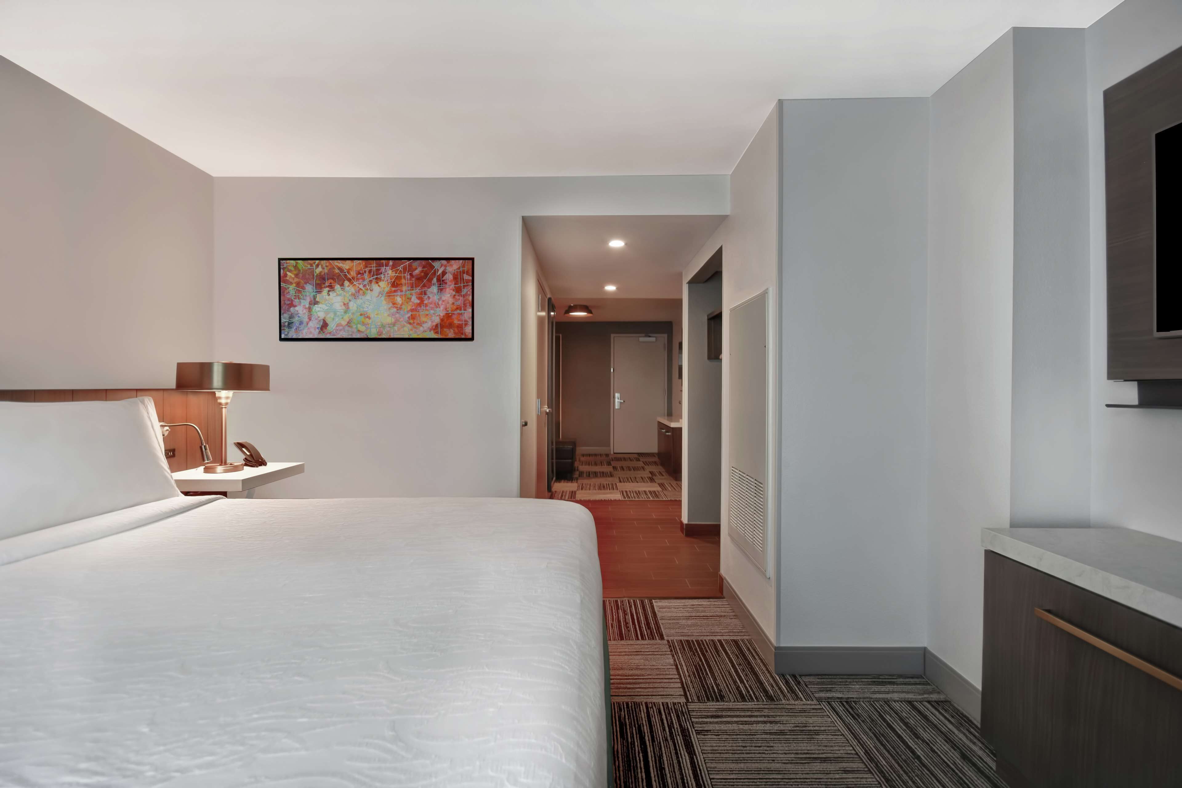 A hotel room with a large bed, a desk with a lamp, a painting on the wall, and a view into the hallway.