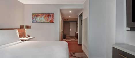 A hotel room with a large bed, a desk with a lamp, a painting on the wall, and a view into the hallway.