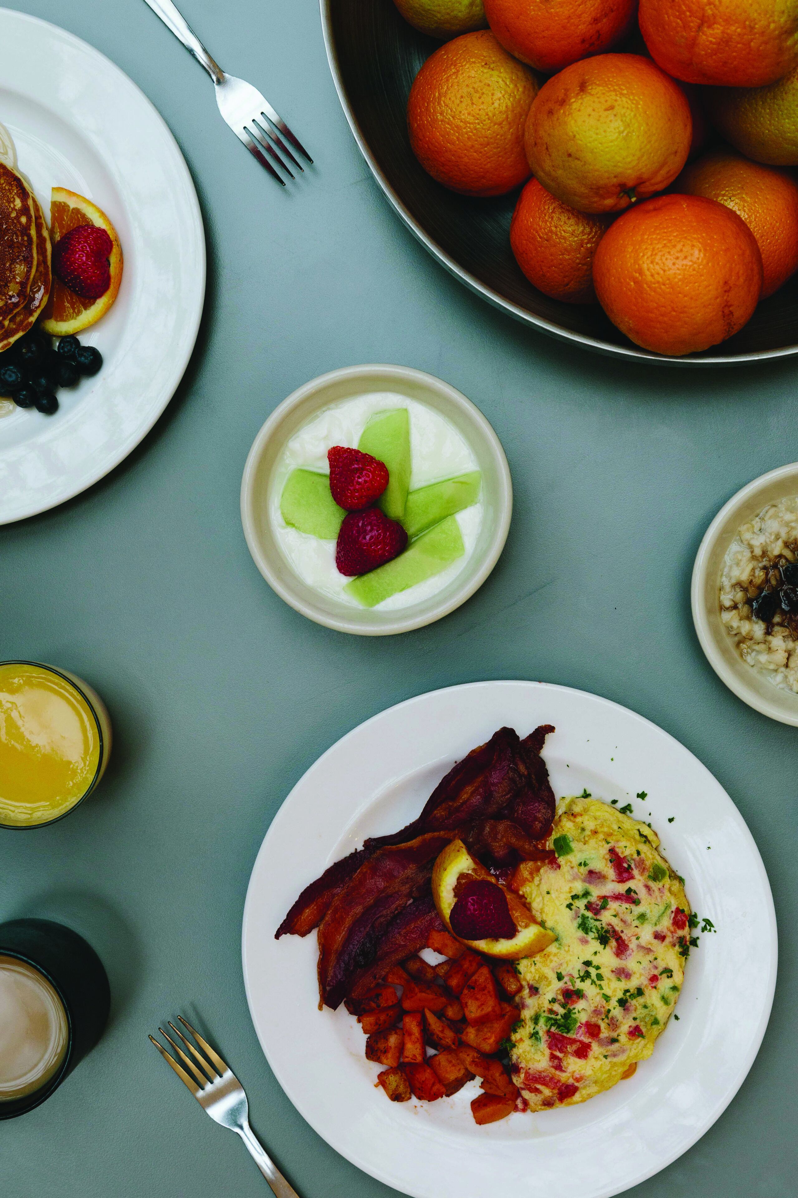 A breakfast table with plates of food, a bowl of oranges, and a cup of coffee.