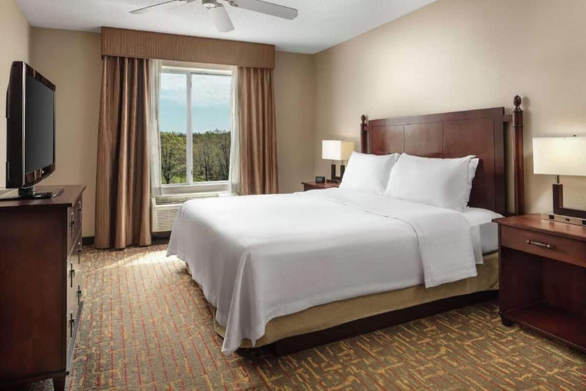 Een hotelkamer met een bed, een televisie, een raam met gordijnen, een plafondventilator en een nachtkastje met een lamp.