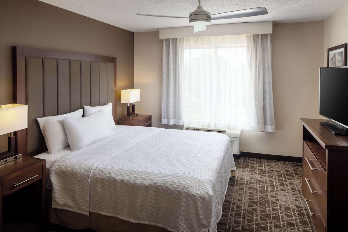 Een hotelkamer met een groot bed, een plafondventilator, een televisie, een nachtkastje met een lamp en een raam met gordijnen.
