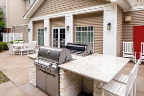Una cocina exterior con parrilla y estufa empotrada, sillas blancas y una puerta roja.