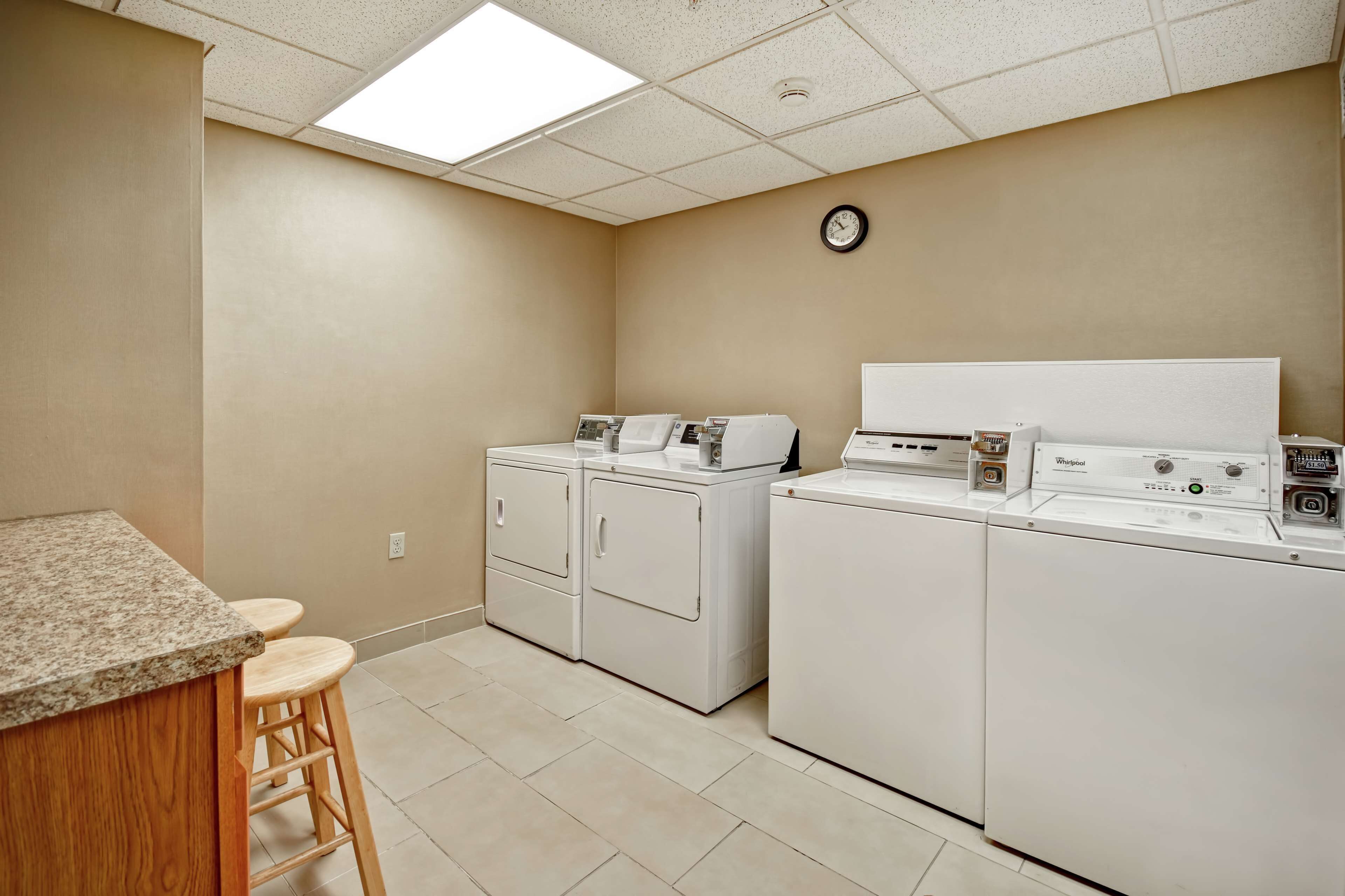 A laundry room with two stacked washers and dryers, a wooden bar stool, and a clock on the wall.