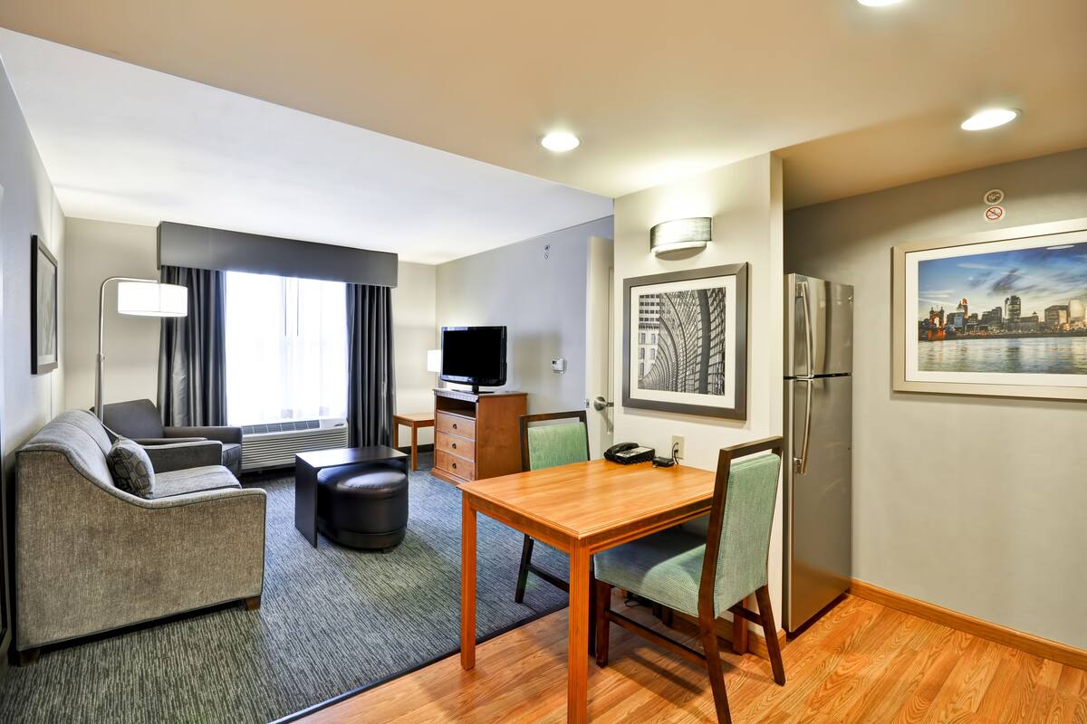 Habitación de hotel compacta con sofá, una pequeña mesa de comedor, televisor, refrigerador y un cuadro con vista a la ciudad.