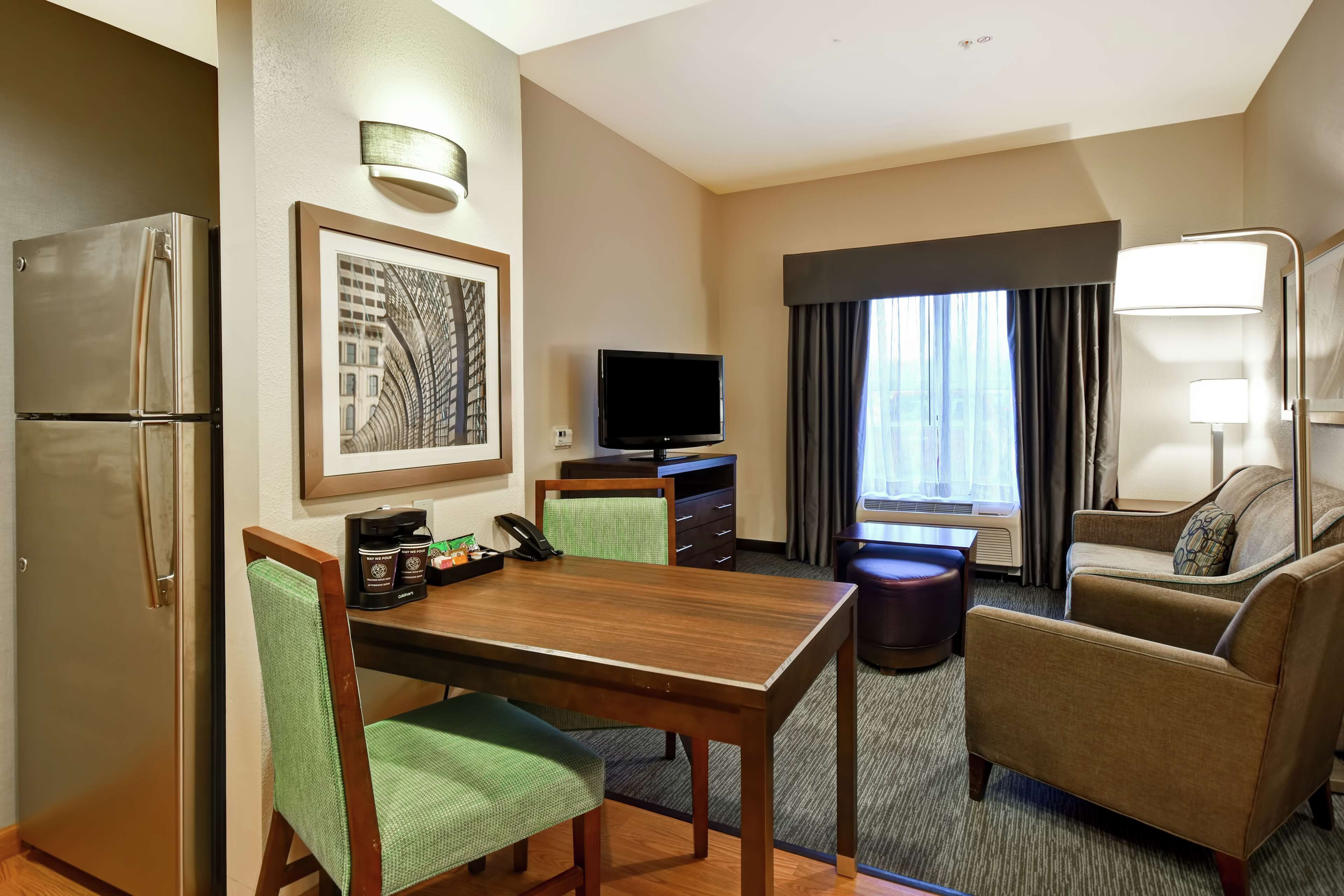 A hotel room with a dining table, chairs, a refrigerator, a television, a coffee maker, a sofa, and a window with curtains.