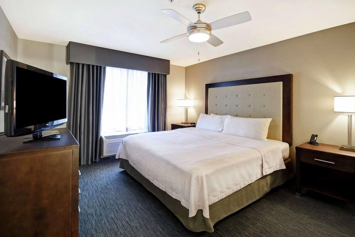 Una habitación de hotel con cama, televisor, ventilador de techo, mesita de noche, lámpara y ventana con cortinas.