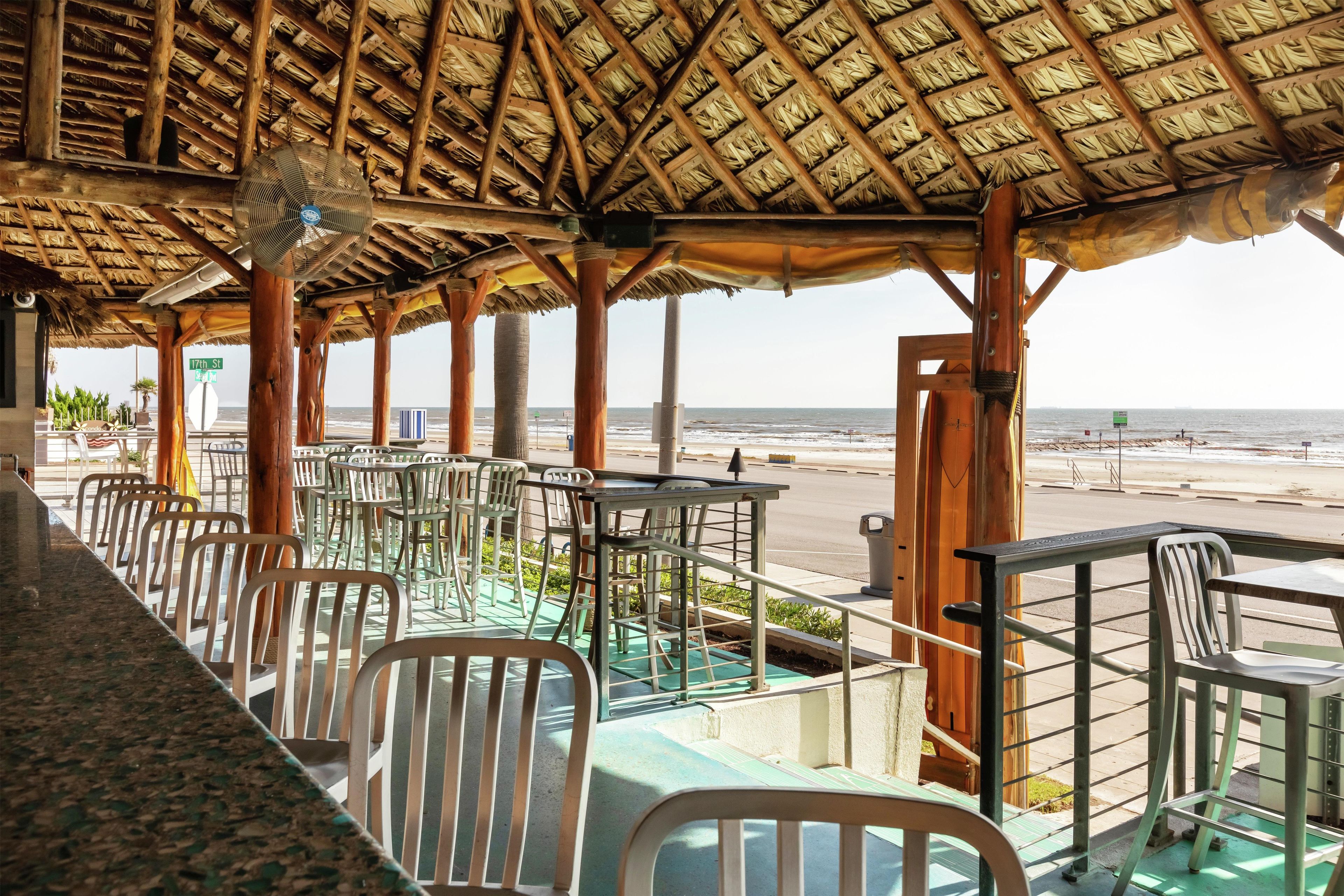 A beachside restaurant with outdoor seating, a thatched roof, and a view of the ocean.