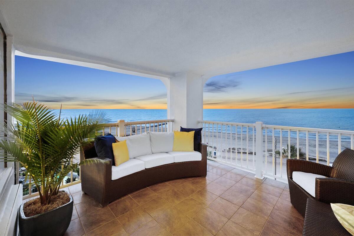 Een balkon met een witte reling, een bruine rieten bank met kussens, een bloempot en uitzicht op de oceaan bij zonsondergang.