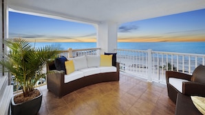 A balcony with a white railing, a brown wicker sofa with cushions, a potted plant, and a view of the ocean at sunset.