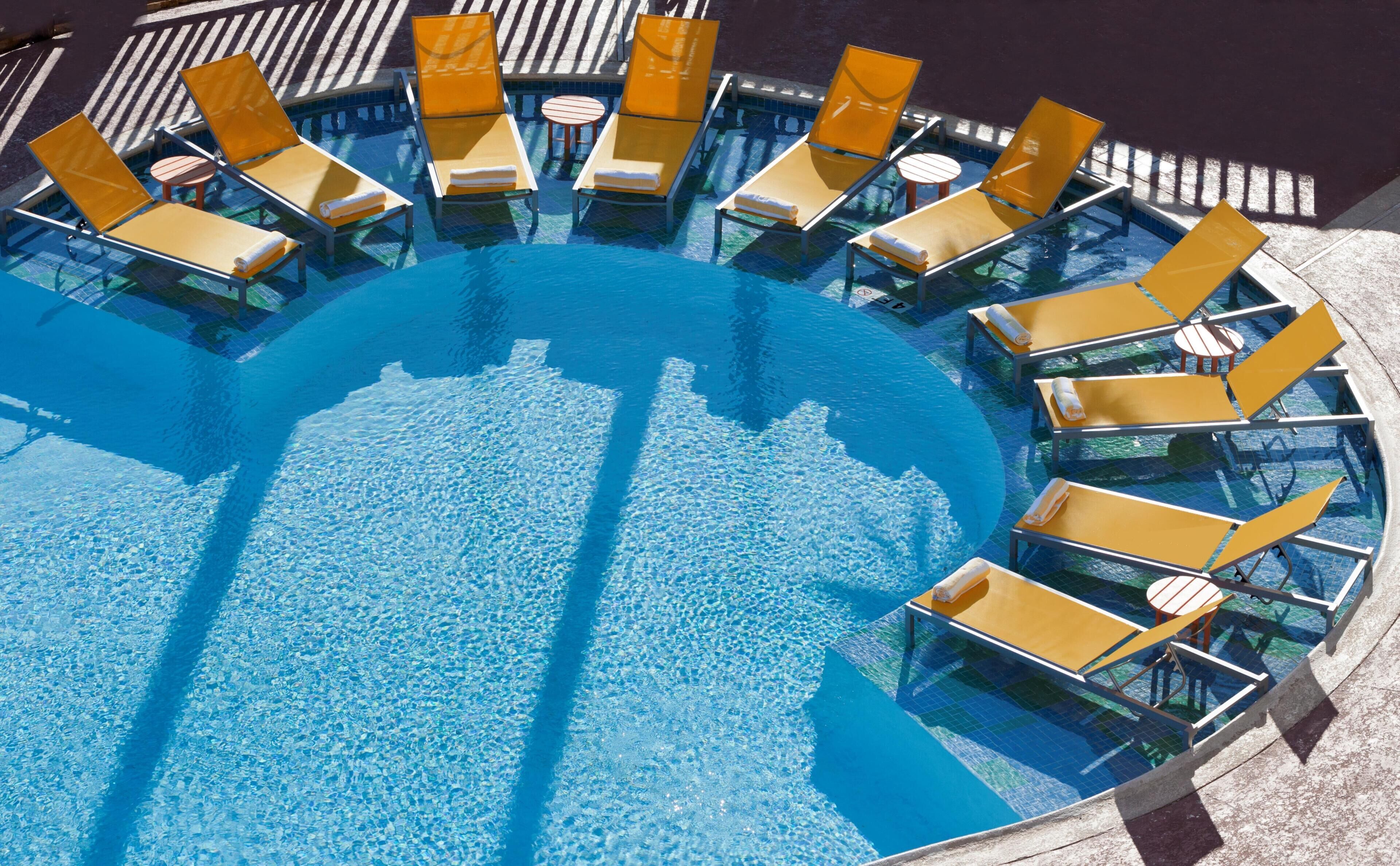 Una piscina circular con agua azul cristalina, rodeada de sillas de descanso amarillas y pequeñas mesas redondas.