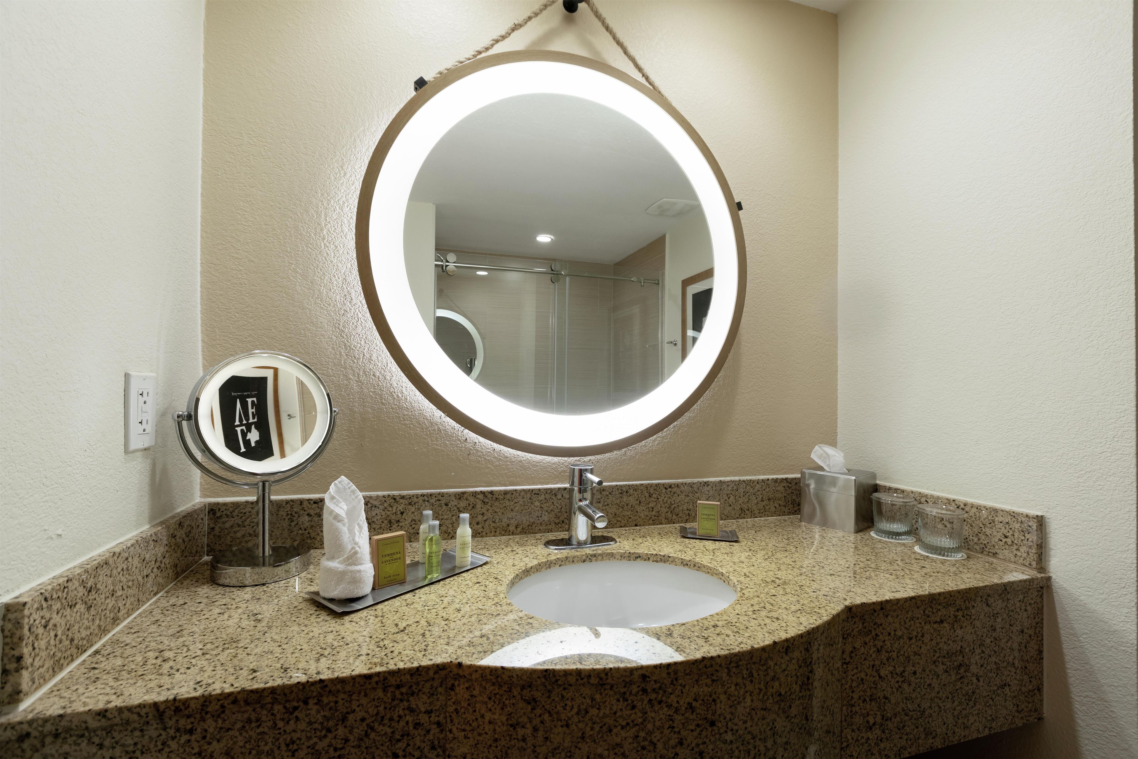 A bathroom with a granite countertop, a round mirror with a light, a sink, and a glass shower enclosure.