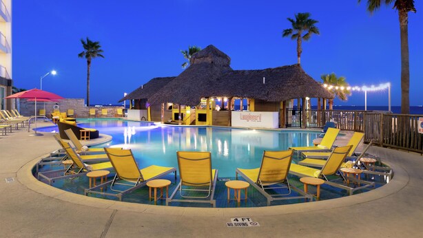 A pool area with lounge chairs, a thatched-roof structure, palm trees, and a sign for 'Longboard Pool Bar'.