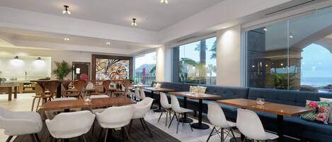 Un espace de repas moderne, avec des tables et des chaises, un canapé et de grandes fenêtres donnant sur l’océan.