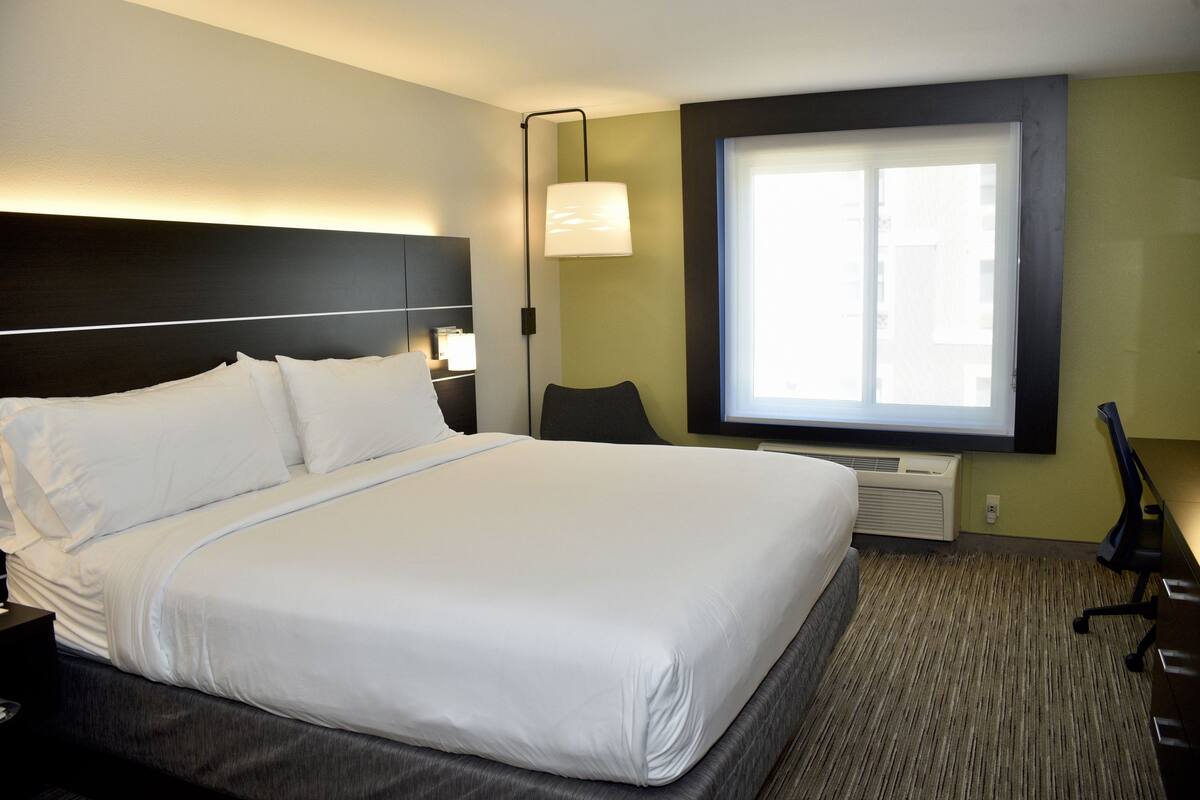A hotel room with a large bed, a desk, a chair, a lamp, and a window.