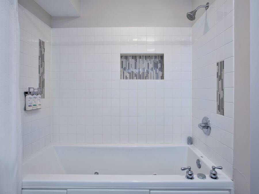 Une salle de bain avec une baignoire blanche, une pomme de douche et une petite fenêtre ornée d’un motif en mosaïque.