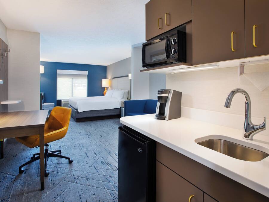 Habitación de hotel compacta con kitchenette, una cama frente a una pared azul decorativa, un escritorio con silla y un baño pequeño.