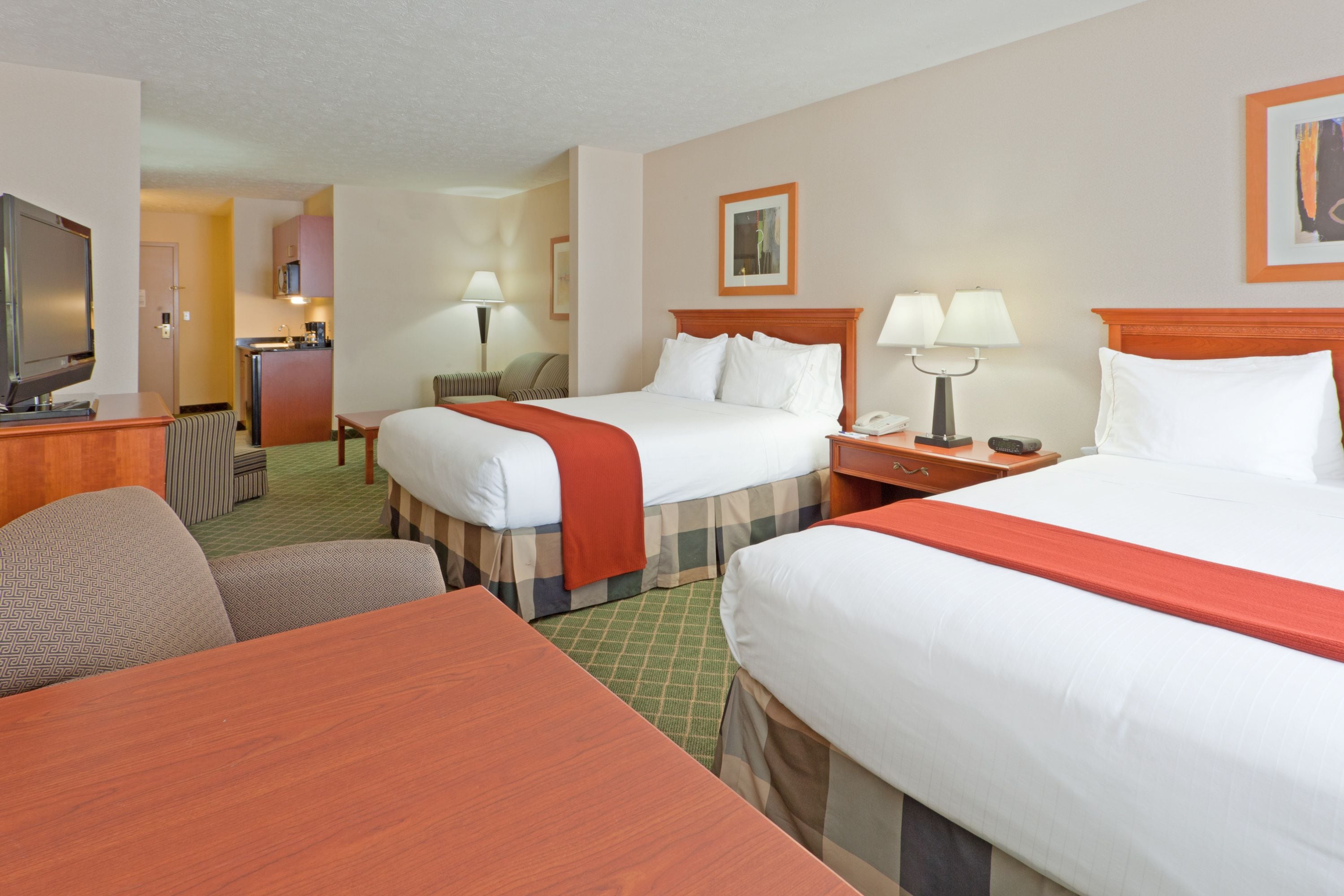 A hotel room with two beds, a desk, a chair, a TV, a lamp, and a small kitchenette.