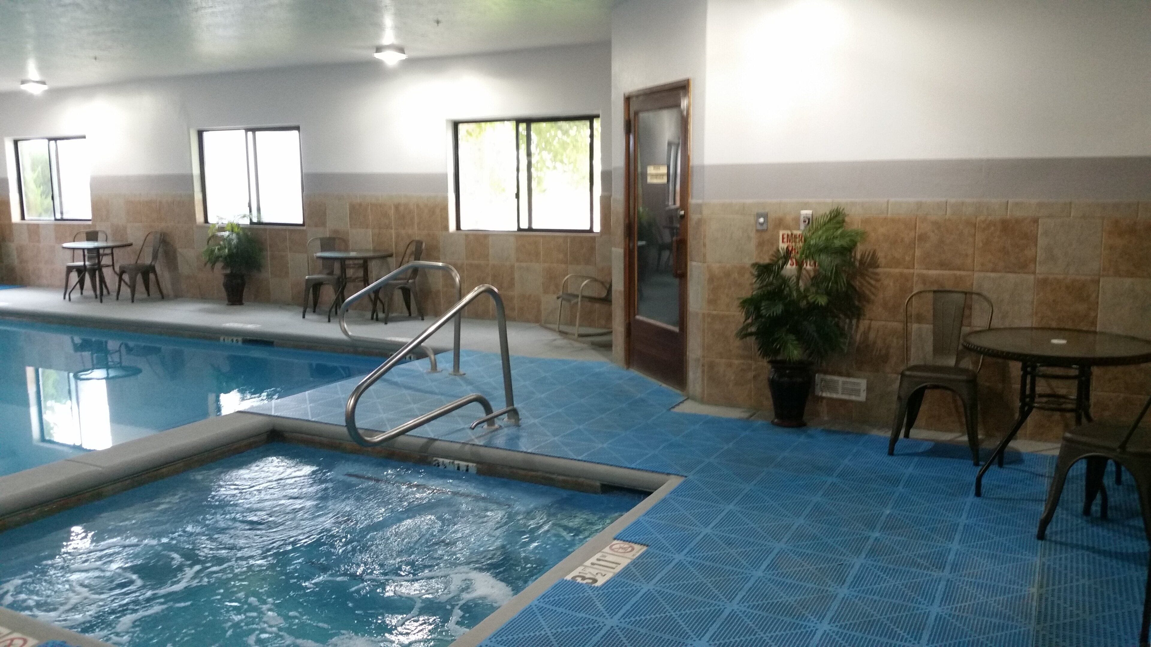 An indoor swimming pool with a blue tiled floor, metal pool ladder, and several tables and chairs around the pool area.