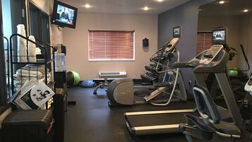 Un gimnasio con cintas de correr, una bicicleta estática y una máquina de remo. Hay pelotas de ejercicio y mancuernas en estantes. Un televisor está montado en la pared.