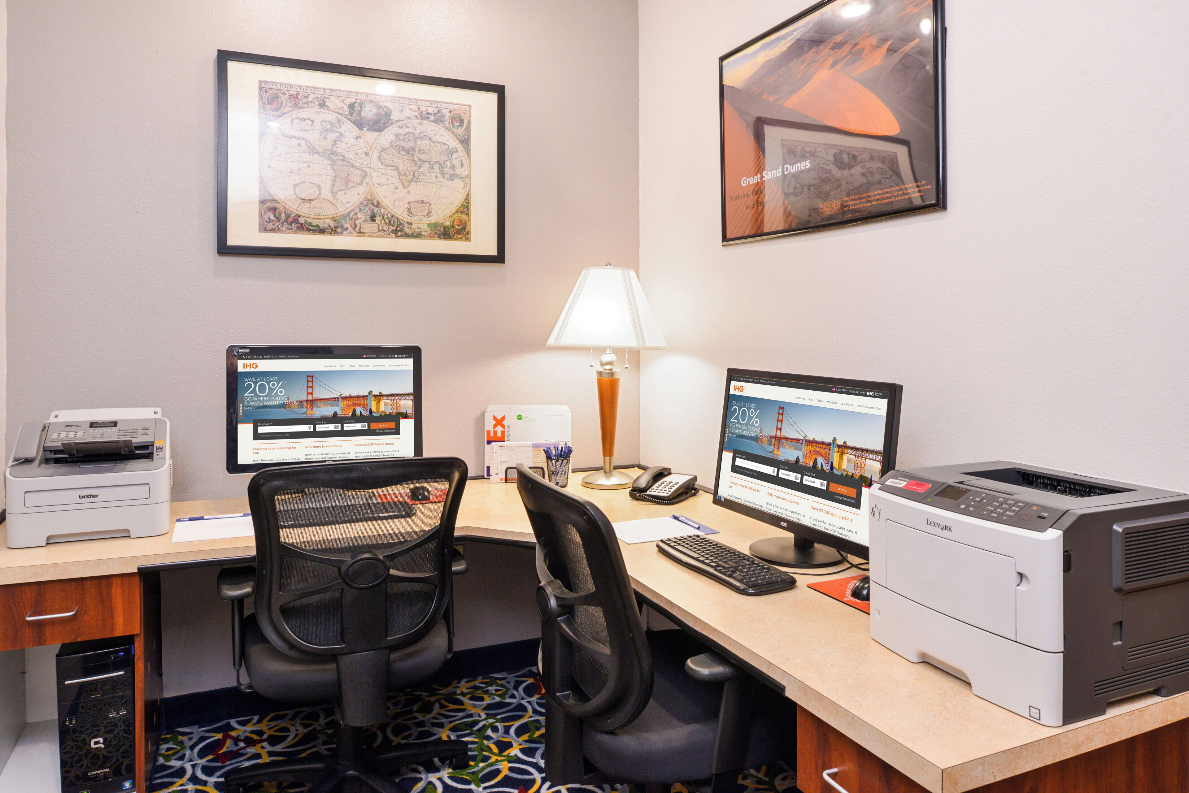 A hotel office with two desktop computers, a printer, a desk lamp, and a framed map on the wall.