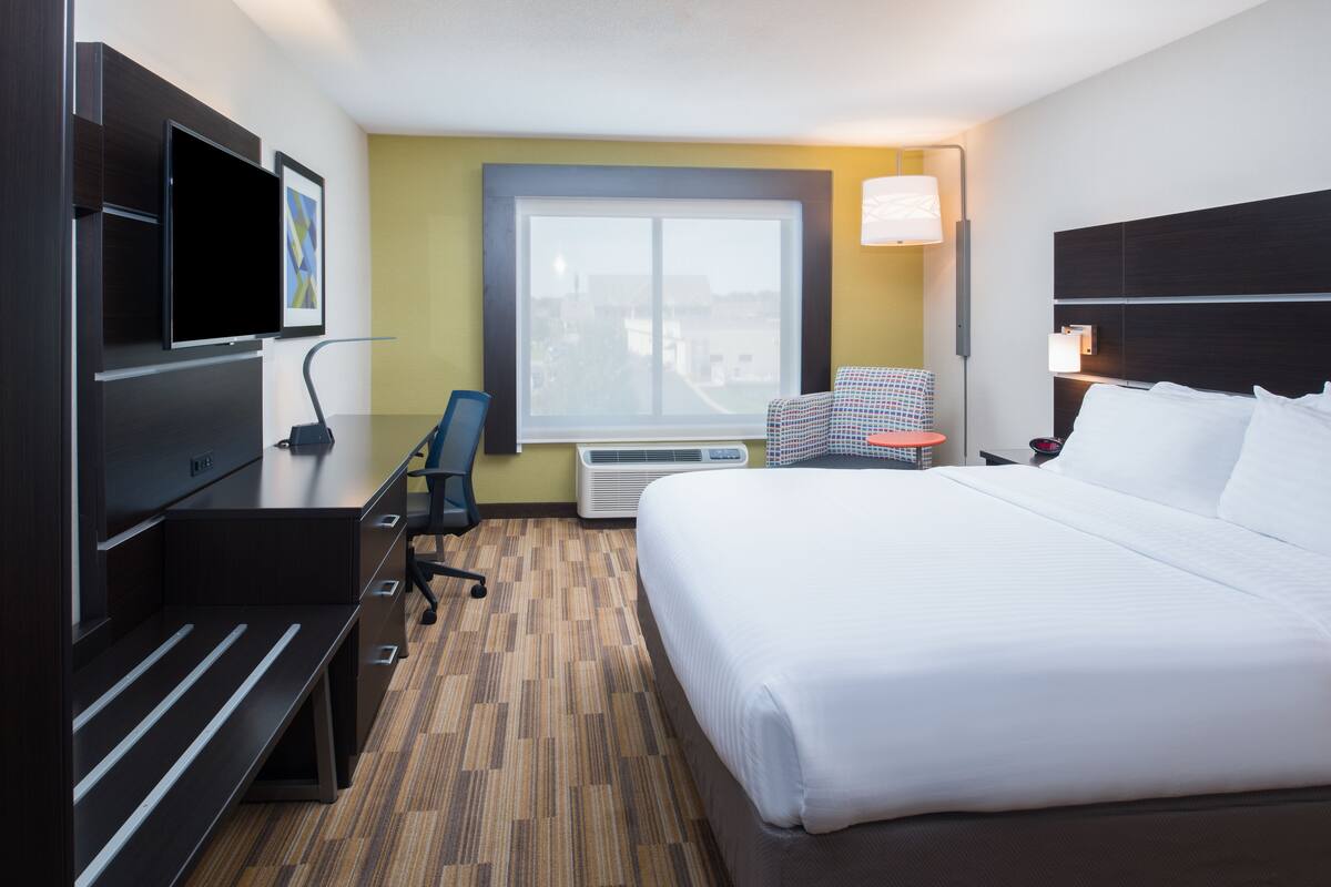 A hotel room with a large bed, a desk with a chair, a TV, a window, and an air conditioning unit.