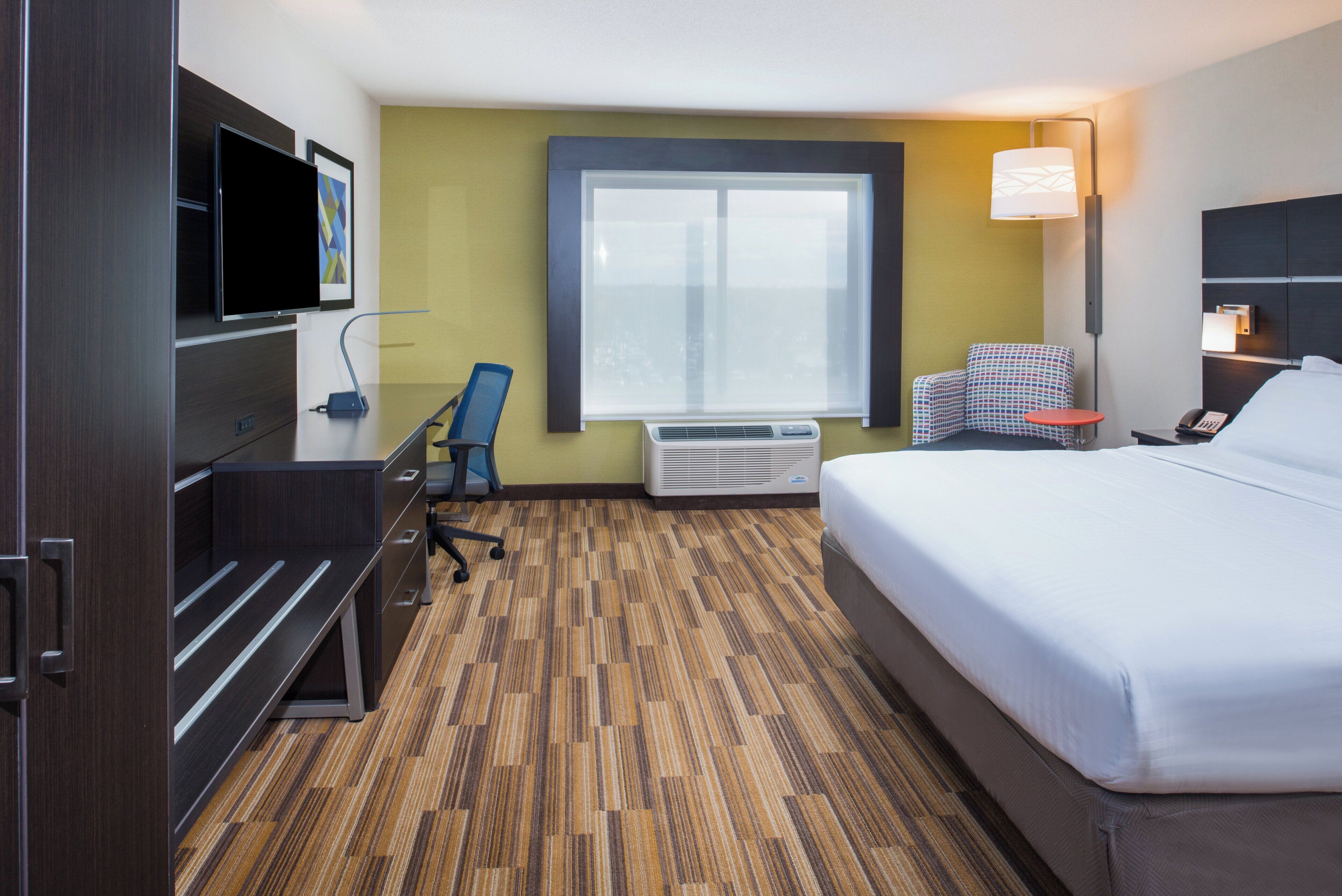 A hotel room with a bed, desk, chair, TV, air conditioning unit, and a window.