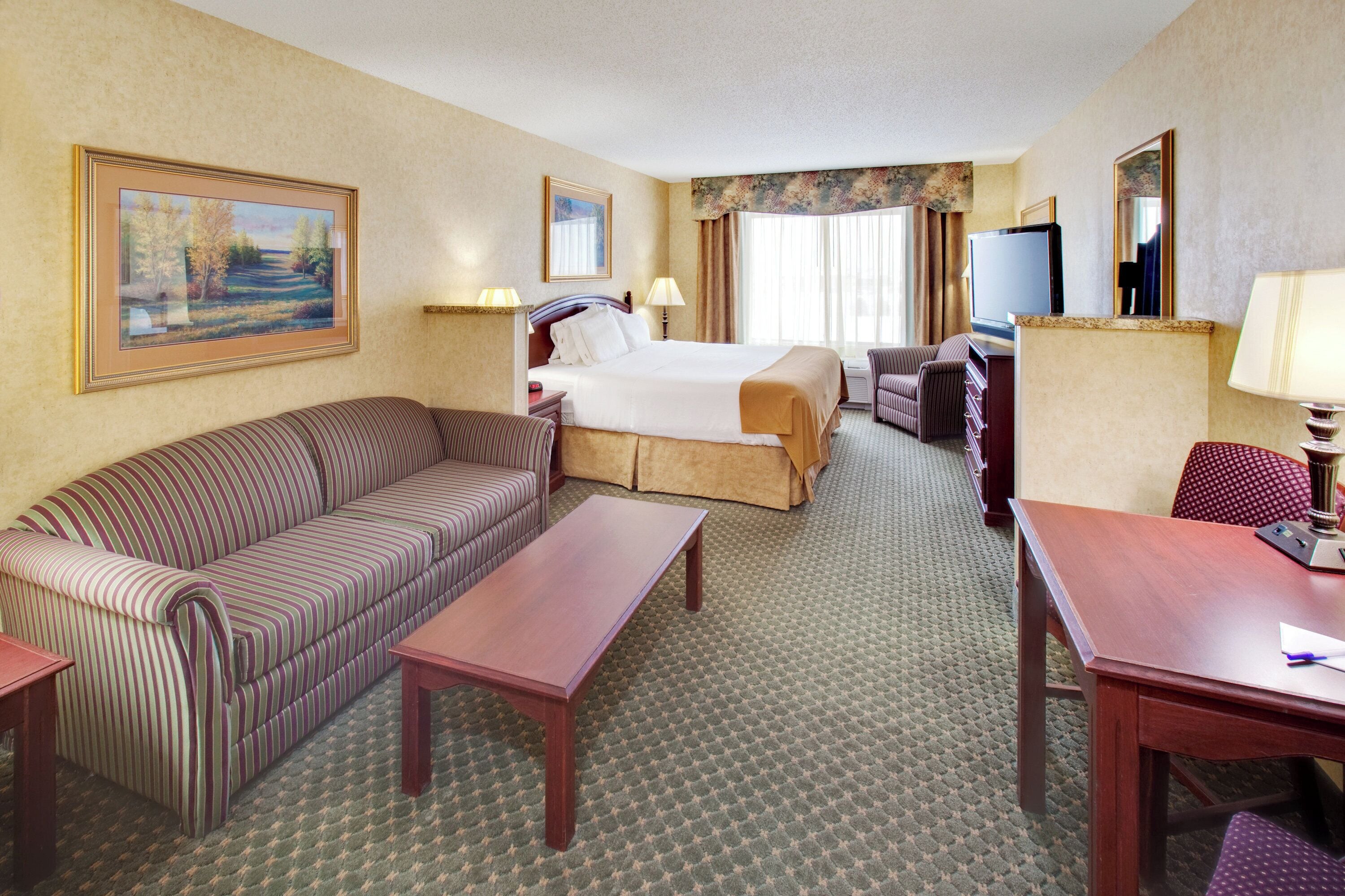 A hotel room with a bed, a sofa, a desk, a TV, and a painting on the wall.